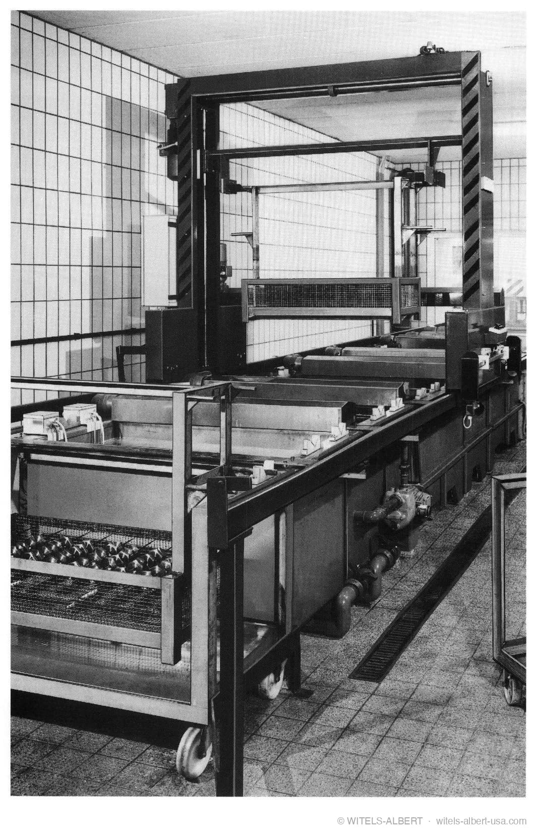 Photograph of an industrial burnishing plant with a gantry hoist above a row of open treatment tanks and a wire basket of parts in a tiled room.