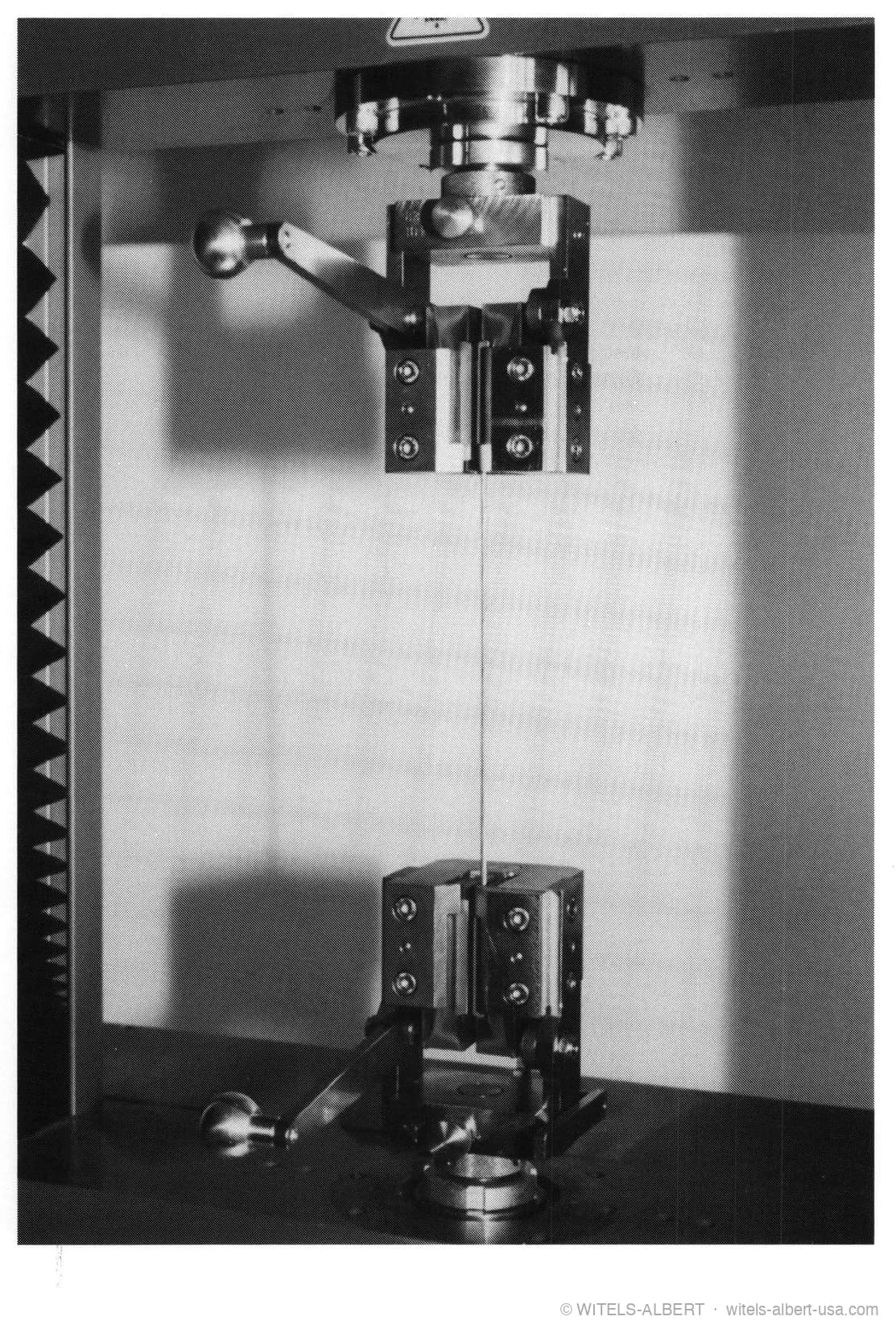 Photograph of a wire specimen clamped vertically between the upper and lower grips of a tensile testing machine.