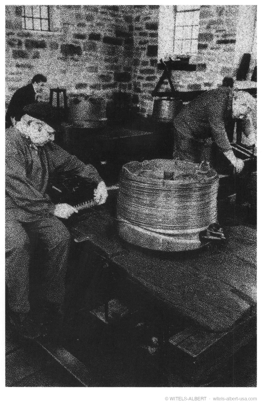 Historic photograph of wire drawers at work in a brick-walled workshop, one man handling a large coil of wire on a bench.