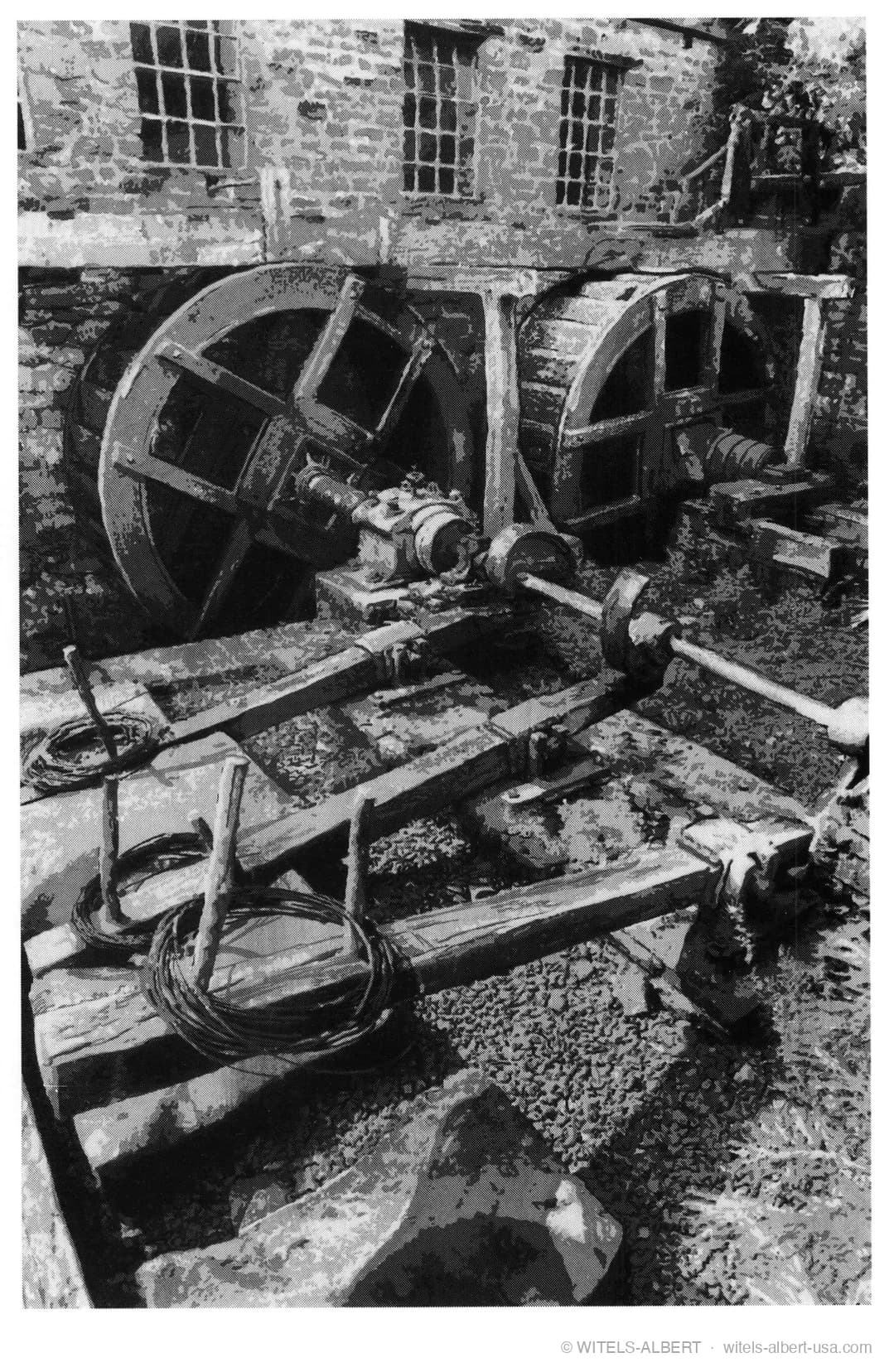 Photograph of historic water-powered wire-washing equipment by a mill wall, with wooden rockers, shafts and coils of wire.